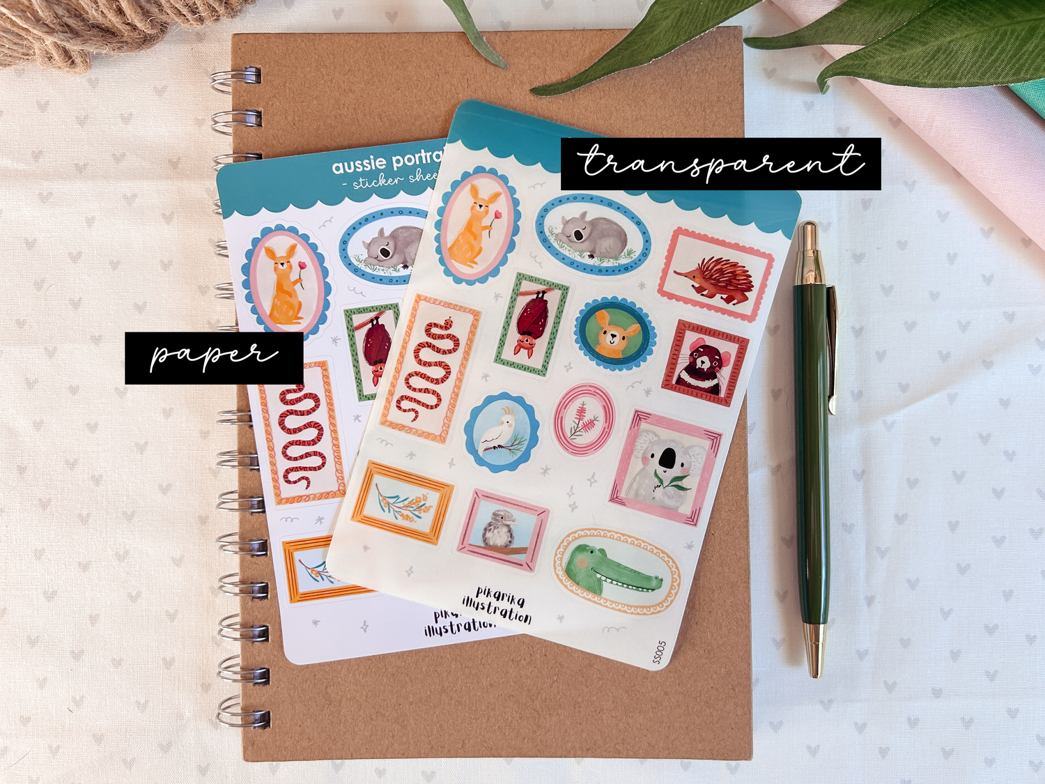 A sticker sheet with various Australian animal illustrations placed on a notebook, with a pen and decorative elements in the surrounding area. The stickers are shown in both paper and transparent variations.